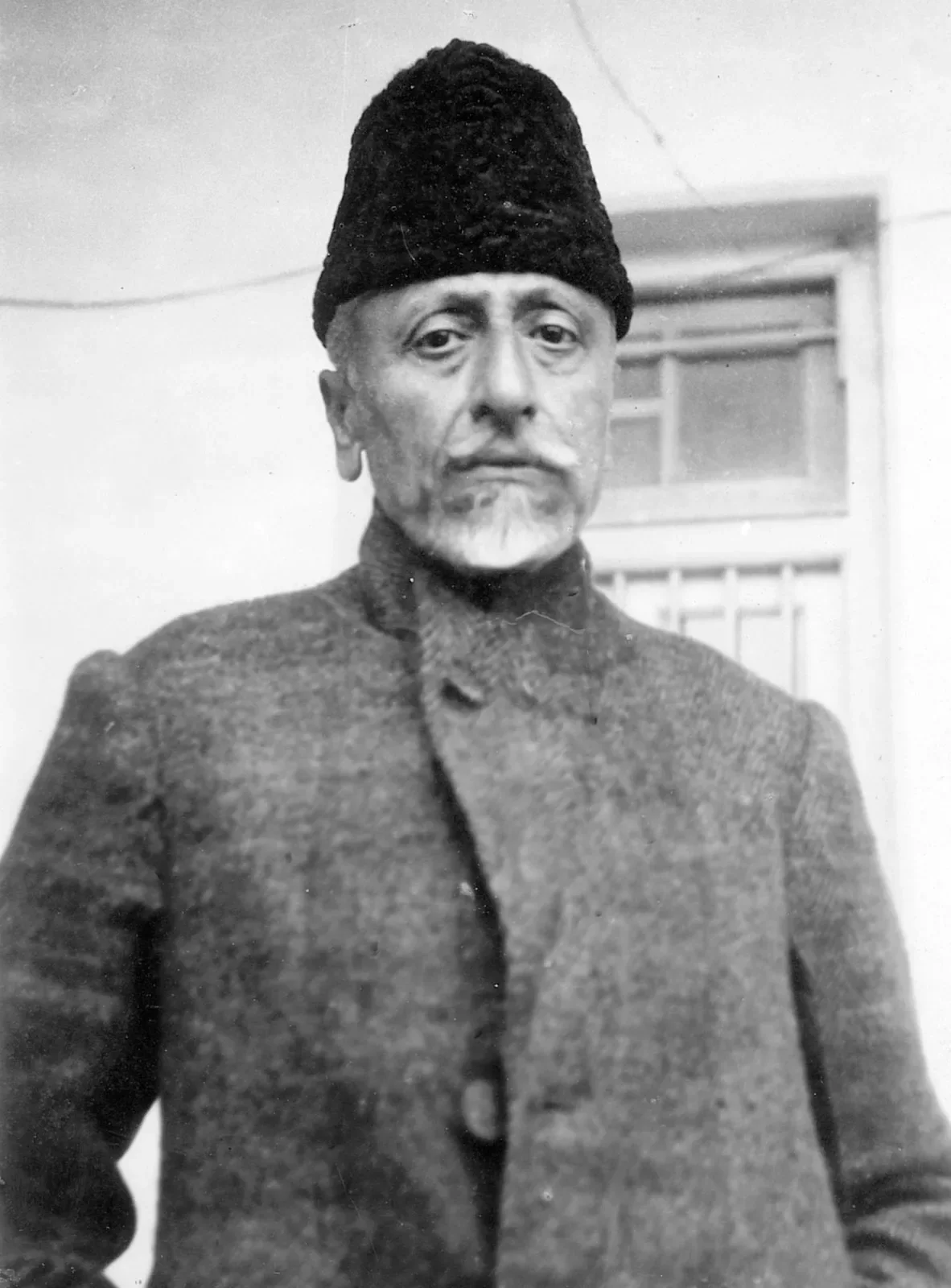 Full-length black-and-white photograph of Maulana Abul Kalam Azad standing in a heavy coat and karakul cap, looking directly at the camera.