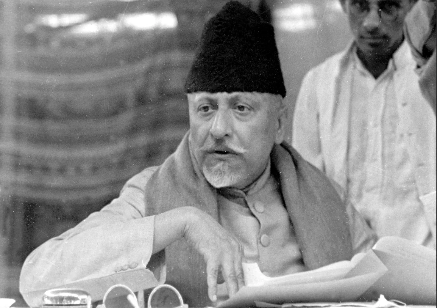 Maulana Abul Kalam Azad seated at a table with papers and spectacles in front of him, speaking, with other men standing behind.